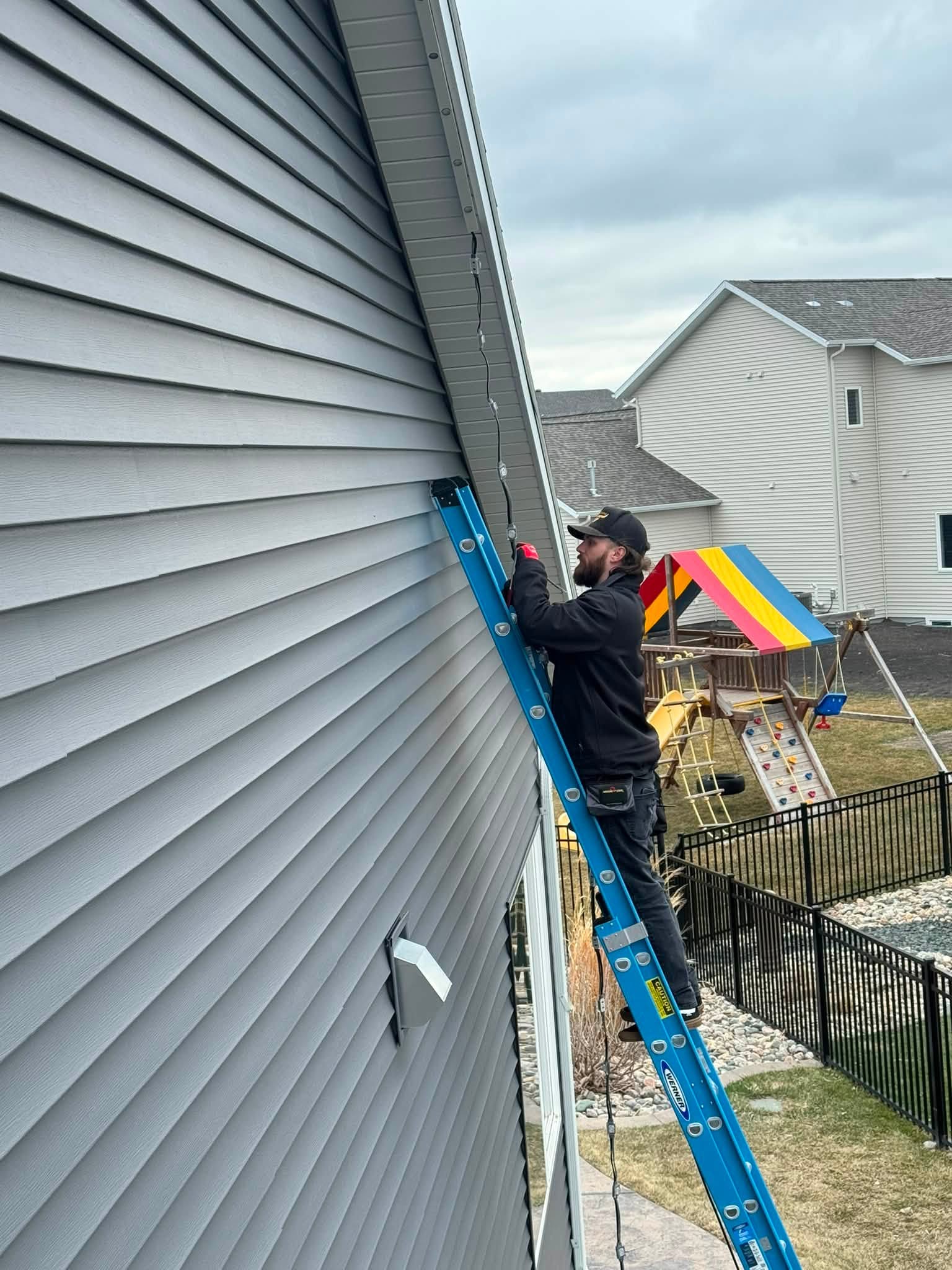 A TiDAL-X installer on a ladder wiring the eave of a grey house by day